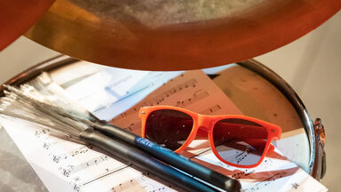 Foto eines Schlagzeugs. Auf diesem liegen ein Notenblatt, eine Sonnenbrille und zwei Drumsticks.