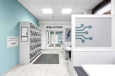 Foto des Flurs vor dem Eingang zur Bibliothek. Dort befinden sich Schließfächer, eine Rückgabebox und Gruppenarbeitsplätze__Photo of the hallway in front of the library entrance. It features lockers, a book return box and desks for group study