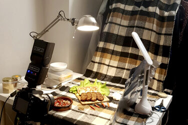 A camera and lamps are pointed at a table on which food is draped for photographing.