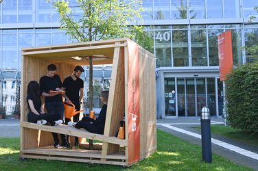 Menschen sitzen in einem großen Holzwürfel vor dem Architekturgebäude der FH Dortmund. Die Würfel sind Teil der Jubiläums-Kampagne.