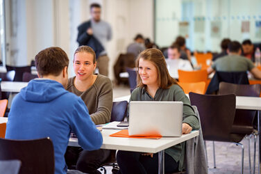 Foto von drei Studierenden, die an einem Tisch sitzen und sich austauschen. Im Hintergrund weitere Studierende an Tischen.__One female and two male students sit together at a table and study. In the background more students at tables.