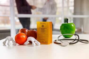 Photo of a still life of various health management merchandise, such as the anti-stress ball. Otherwise, vaccination card, stethoscope and apples.