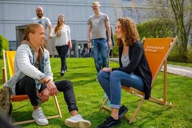 Foto von einer Studentin und einem Studenten in Liegestühlen mit der Aufschrift Fachhochschule Dortmund auf einer Wiese auf dem Campus. Sie unterhalten sich, weitere Studierende nähern sich.__Two students sit in deckchairs with the label Fachhochschule Dortmund on it on the campus and chat. Other students in the background.