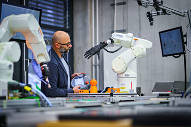 Photo of a laboratory employee standing on the conveyor belt between two robot gripper arms and holding a remote control in his hand.