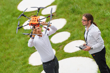 Photo with two men. One is holding a drone high above his head and looking up at it. The other is holding the remote control in his hand and is also looking at the drone.