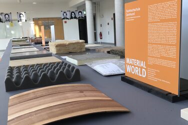 View into the foyer of the Faculty of Architecture. There is a small information board about the "Material World" exhibition on a table. In front of it, various material samples are lined up next to each other.