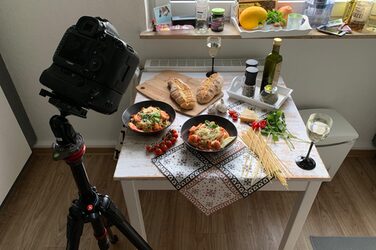 A camera on a tripod at the front left is pointed at a table, on it two plates with a pasta dish, surrounded by corresponding ingredients and spices.