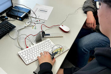 Auf einem Tisch liegen eine Computertastatur und Kabel mit technischen Bausätzen. Rechts daneben sitzen zwei Personen.