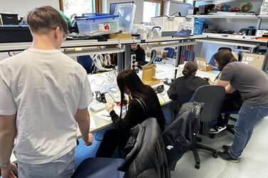 In einem Raum sitzen und stehen mehrere Personen an Tischen, der Raum ist gefüllt mit technischen Gegenständen.