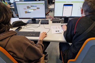 Zwei Personen sitzen an einem Tisch mit zwei Bildschirmen.