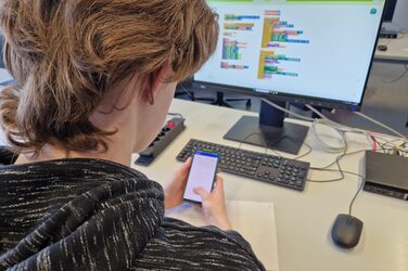 Eine Person sitzt an einem Tisch mit einem Computer und hält ein Smartphone vor sich.