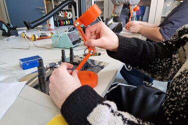 Zwei Hände halten eine Tischlampe, im Hintergrund sind technische Geräte zu sehen.