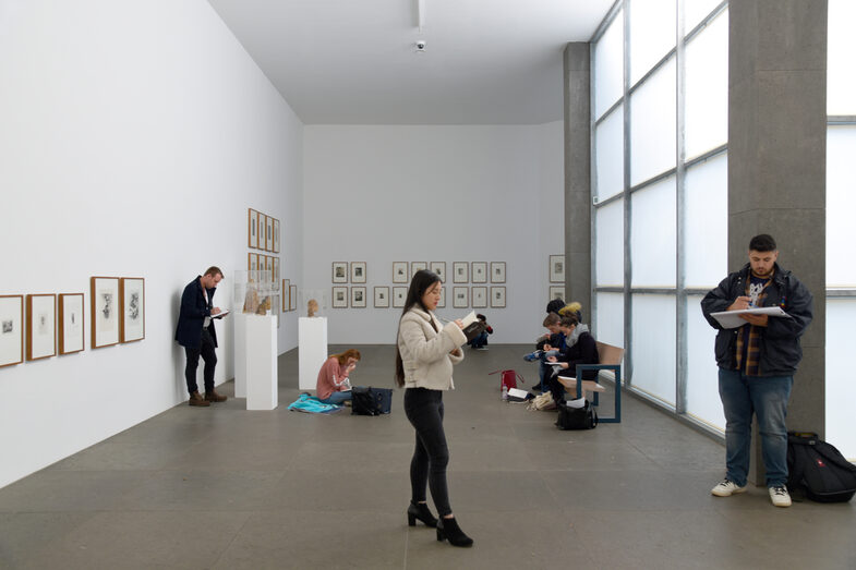 People sit and stand in a gallery, holding pads and pens in their hands.