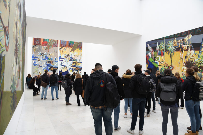 People stand in a gallery. Large paintings hang on the walls.