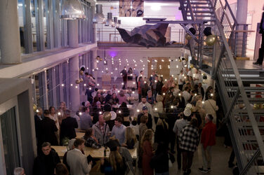 Viele Gäste feiern im festlich beleuchteten Foyer des Fachbereichs mit mehreren Etagen und Lichterketten. Die Stimmung wirkt lebhaft und gesellig.
