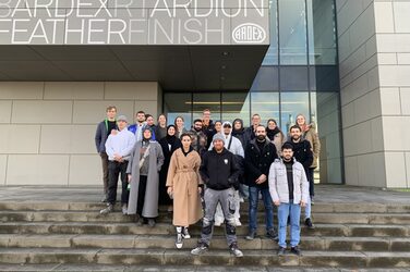 Gruppenfoto mit 21 Personen, welche mittig im Foto auf einer breiten Treppe vor einem sandfarbenen Gebäude mit der Aufschrift "Ardex" stehen. Im Hintergrund sieht man große Fensterflächen des Gebäudes