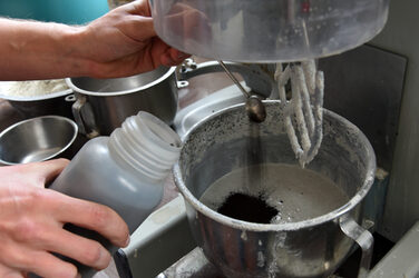 The picture shows a metal bowl in which fresh concrete has just been mixed by machine. The bowl is standing on a table, locked in a device under a stirring rod with which the concrete is being stirred. A small pile of black powder lies on the surface of the liquid concrete. Two hands reach in from the left edge of the picture, one holding an open plastic bottle and the other using a small dosing spoon to sprinkle black pigment powder into the concrete mixture from above for coloring. Further metal bowls can be seen in the background on the left-hand side of the picture.