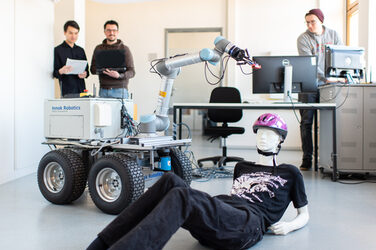Photo of a rescue robot standing next to a life-size dummy on the floor. Behind it on the left are two men with documents looking at the robot. At the back right is a man standing at a table looking into the computer.