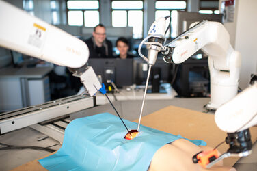 Surgical robot with endoscopes and plastic torso in the foreground. The endoscopes are inserted into an opening in the torso. Behind them, two employees at the computer workstation can be seen out of focus.