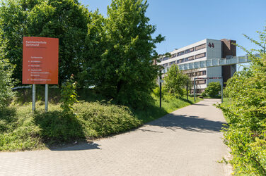 Photo of the sidewalk leading to the building Emil-Figge-STraße 44 of the Fachhochschule Dortmund. To the right of the sidewalk, a signpost with the Fachhochschule Dortmund logo.