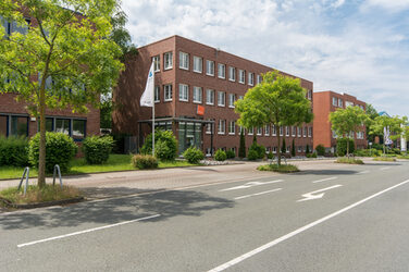 Photo of the Fachhochschule Dortmund site in Otto-Hahn-Straße from the outside. It is a red brick building. In the foreground are a road and some trees.