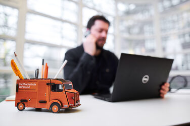 Photo of a pencil box in the form of a car on a desk. Out of focus behind it, a man sits at his laptop and makes a phone call __Photo of a pencil box in the form of a car on a desk. Out of focus behind it, a man sits at his laptop and makes a phone call.