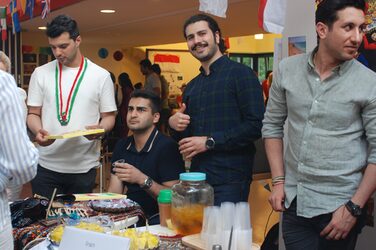 Four students stand around a decorated table with food at the culture stand. One student looks into the camera and smiles.