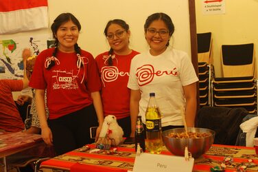 Kulturstand Peru: 3 Studentinnen stehen hinter einem dekorierten Tisch und lächeln in die Kamera.