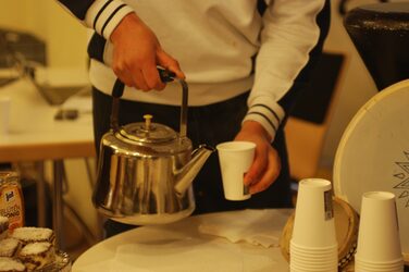 A person pours something from a metal teapot into a paper cup.