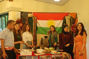 Six people stand around the decorated table at the Kurdish cultural stand and look into the camera.