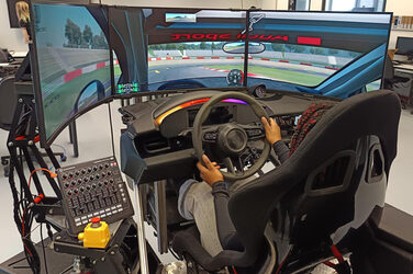 Eine Person steuert sitzend das Lenkrad in einem Fahrsimulator mit drei Bildschirmen.