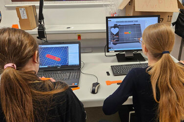Zwei Personen sitzen an einem Tisch, vor ihnen stehen Computer, deren Bildschirme jeweils ein Modell für Schlüsselanhänger zeigen.