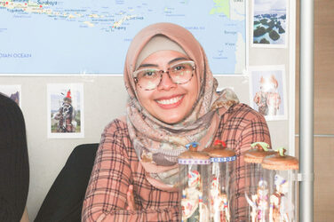 Eine der Ausstellerinnen beim "Marktplatz der Möglichkeiten" der Internationalen Woche sitzt lächelnd an einem Tisch mit Flyern. Im Hintergrund ist eine Landkarte__One of the exhibitors at the International Week's "Marketplace of Opportunities" sits smiling at a table with flyers. In the background is a map