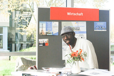 Ein weiterer Ausstellertisch des "Marktplatzes der Möglichkeiten": ein junger Mann sitzt am Messestand und lächelt in die Kamera__ Another exhibitor table of the "Marketplace of Opportunities": a young man sits at the booth and smiles into the camera