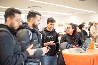 Vier Studierende spielen an einem orangefarbenen Stehtisch ein Spiel an einem Laptop und lachen dabei. Four students are playing a game on a laptop at an orange high table and laughing.