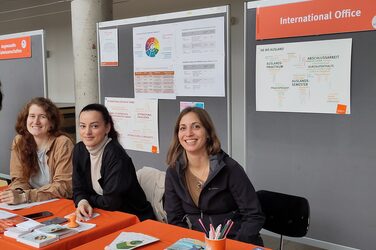 Drei junge Frauen sitzen an den Messeständen des international Day und freuen sich auf interessierte Studierende. Three young women sit at the International Day stands and look forward to meeting interested students.