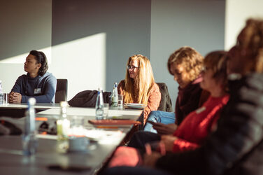 Fünf Personen an einem Konferenztisch blicken interessiert nach links.Die Sonne scheint in den Raum__Five people at a conference table look to the left with interest. The sun shines into the room