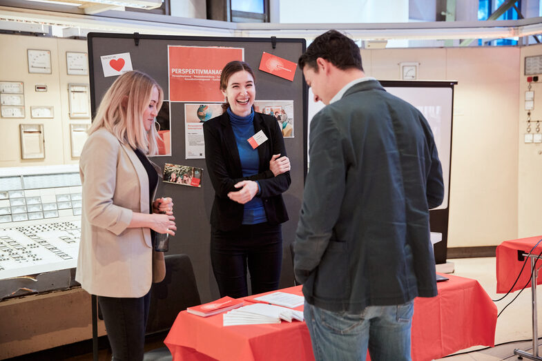 Zwei Hochschul-Mitarbeiterinnen am Info-Tisch des Perspektivmanagemets in der DASA im Gespräch mit einem Besucher.__Two female university employees in conversation with a visitor at an info table at the event venue DASA.