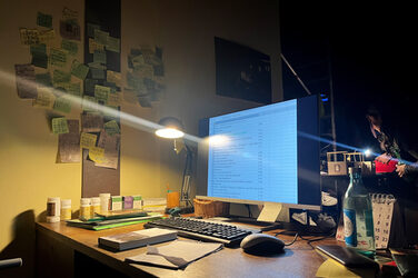 View of a desk illuminated only by a desk lamp: You can see a switched-on screen with keyboard and mouse, several tins of medication, pads, a calendar and other typical utensils. There are lots of Post-It notes written on the wall.