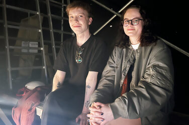 Zwei Personen sitzen auf einer Art Gerüstlauffläche mit Leitern im dunklen Hintergrund und blicken freundlich in die Kamera.