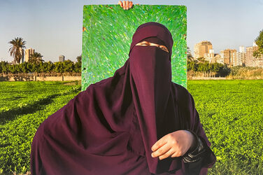 A person wearing a wine-red nikab and a robe of the same color is standing in front of a colorful, abstract painting that someone is holding up right behind her. A meadow can be seen around it, with palm trees and beige-colored skyscrapers in the background.