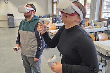 Zwei Personen stehen in einem Seminarraum, sie haben jeweils einen VR-Brille vor den Augen und halten Steuerungselemente in den Händen.