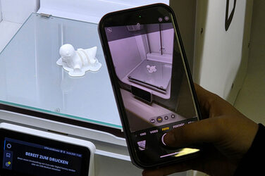Eine Hand hält ein Smartphone, um ein Foto von einer Miniaturfigur zu machen, die in einem 3D-Drucker liegt.