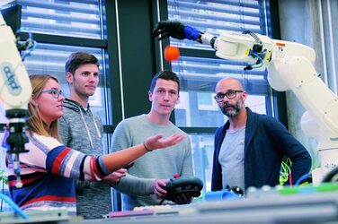Foto von vier Personen, die um eine bionische Hand an einem Industrieroboter herumstehen. Diese Roboterhand lässt einen Ball fallen, den eine junge Frau auffangen will.