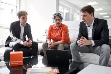 Photo of three people sitting next to each other on a couch. There is a notebook on the table and the three people are looking at the screen of the notebook.