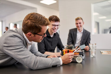 Photo of three people at a table on which a 4-wheeled robot is standing. One of the people is leaning over the table so that he can look at the back of the robot. The other two people watch attentively and pay attention to what the other person wants to show them.