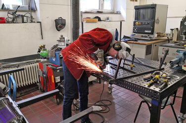 In a workshop, a person bends over a table and works on a metal frame with a grinder, sparks flying.