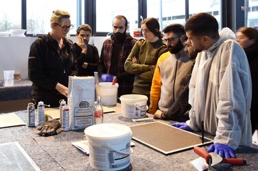 Sechs Studierende stehen an einem mit Malervlies abgedecktem Tisch auf dem Produkte wie Dekorspachtelmasse der Marke Ardex stehen. Außerdem liegt eine unbehandelte Mustertafel auf dem Tisch.