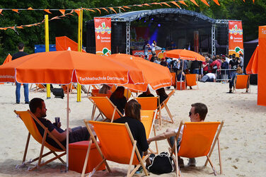Menschen sitzen auf orangefarbenen Sonnenstühlen unter orangefarbenen Sonnenschirmen. Im Hintergrund ist eine Bühne zu sehen.