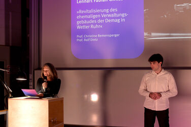 Dekanin Prof. diana Reichle steht am Rednerpult und liest, während ein Student daneben auf der Bühne steht. Hinter ihnen zeigt die Präsentationsfolie ein Projekt mit Titel und Namen in violettem Licht.